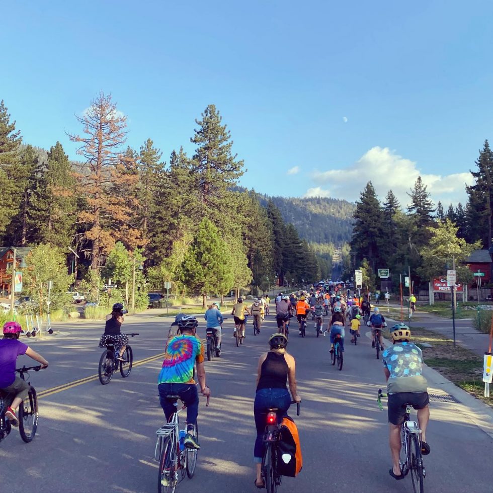 A large group of cyclists riding down a street with trees and mountains in the background.